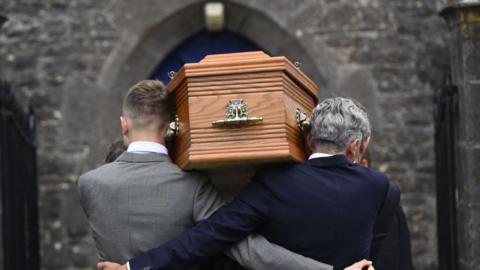 The brown coffin of Lord Mount Charles, being carried at St Patrick's Church of Ireland in Slane. On the left is a man with fair hair and a grey suit, on the right, a man with grey hair and a dark blue suit.