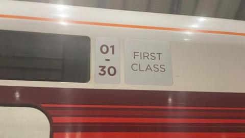 The outside of a train with a sign saying 'First Class' on it. The train has a white top half and red stripy bottom half.
