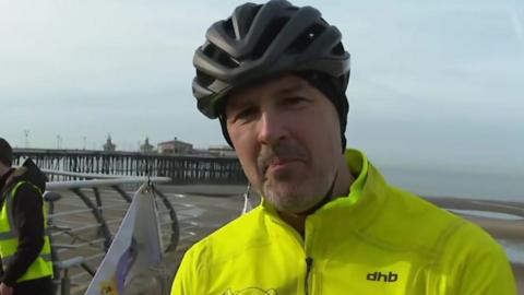 Paddy McGuinness, in a black cycle helmet and a bright yellow-green jacket, stands in front of the beach at Blackpool, with a pier in the background