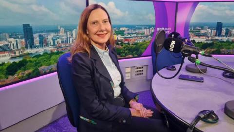 Deborah Cole sat in a BBC Radio Solent radio studio with purple branding around her and a artificial background showing a city skyline.