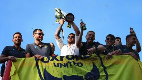 Oxford United boss Des Buckingham lifts the League One play-off promotion trophy on a bus in Oxford