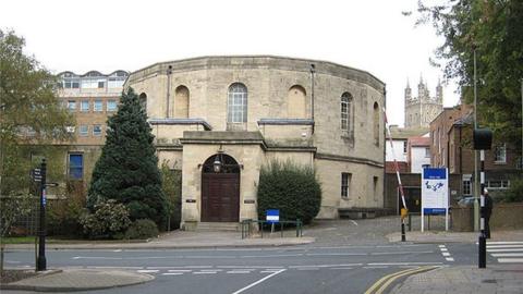 The entrance to Gloucester Crown Court. The building is made of stone brocks and is circular with a large dark brown wooden door.