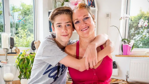 Alison Lapper cuddles her son Parys at their home in 2014. Alison is wearing a pink dress and a streak of her blonde hair is dyed pink. Parys is wearing a grey t-shirt and grey jeans. Artwork can be seen in the room behind them.