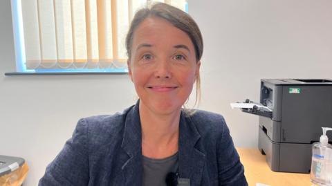 A woman sits in front of a computer in a GP surgery in Grimsby staring at the camera wearing a grey t-shirt and navy blue jacket. The walls are white and there is a window behind her with cream coloured blinds. A large printer is on the desk behind her.