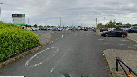 The image shows a car park, with several cars parked in marked spaces, and a central road runs through the lot with an arrow indicating the direction of traffic. On the left side, there is a green hedge and a sign displaying the name of the car park along with other details. The sky is overcast, and lighting poles are scattered throughout the area. 