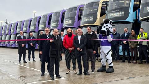 People gathered to cut the ribbon on the new buses which are lined up behind them.