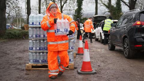 Cars queue up next to large stacks of water bottles. A woman in an orange high vis jacket and trousers is carrying a pack of six bottles. 