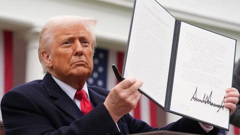 U.S. President Donald Trump displays a signed executive order imposing tariffs on imported goods during a trade announcement event in the Rose Garden at the White House in April. The President is waring a dark blue coat and a red tie. He is holding up the booklet containing the order.