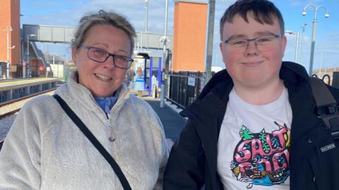 Bradley Watson is standing next to his mother Amanda Watson at Newsham station. He is 20 years old and wearing a blue jacket with white t-shirt. She is wearing a white fleece and they are both smiling.