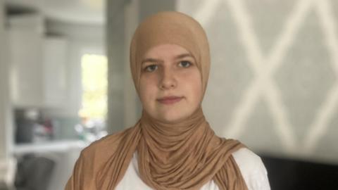 Aleyah Knight. She is pictured in a house wearing a beige hijab and a white t-shirt. She is looking at the camera and smiling