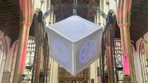 A grey cube, installed between two pillars in a church. Three sides of the cube are visible, and each had a geometric pattern on it. Purple light is shining through this. The pillars are lit up with pink lights. 