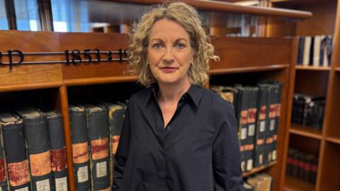 A woman wearing a black dress stands in front of old files 