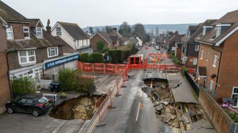 An aerial view of a residential street in Godstone showing a large collapsed section of road with exposed ground, fencing and barriers blocking traffic, with houses and parked car on either side.