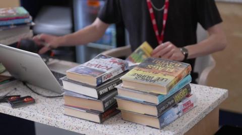 A library worker wearing a black T-shirt checking a pile of eight novels on his silver laptop in a library