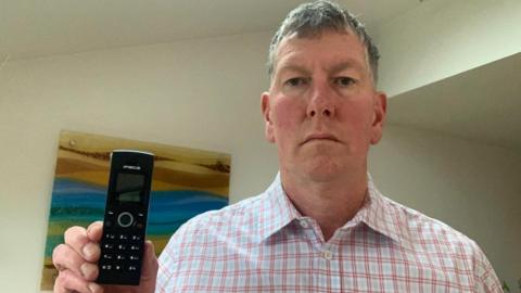 Paul Toplass stands in his office holding a black portable telephone handset. He has short grey hair and wears a white and pink checkered shirt.
