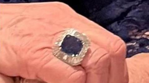 A hand with a large ring on it. The ring is square and has a large purple sapphire stone in the middle and is surrounded by diamonds