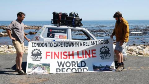 Two men stand next to a small silver car which is parked in front of the sea. The man on the left is standing next to the boot of the car. He is wearing a shirt and short. The man on the right, standing next to the bonnet of the car, is also wearing shorts and a shirt. They are both holding a very large white sign which reads London to Cape Town in a Reliant Robin. Finish Line World Record. 