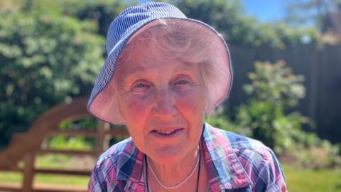 Woman is hat with pink and purple checked shirt with white necklace