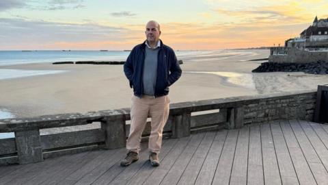 A bald man wearing a navy blue jacket, grey humper and beige trousers stands on a wooden platform with a beach behind him