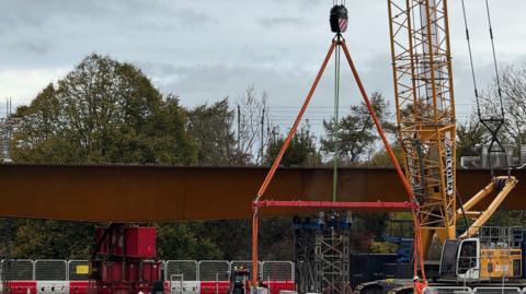 A tall yellow crane is operating next to a large steel bridge. The area on the ground underneath the bridge is blocked off with temporary construction fencing. 