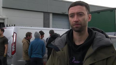 Daniel Sheehy. He has short dark hair and is wearing a green coat over a black hoodie. Behind him are drivers crowded around a silver van.