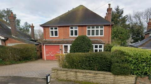 The image shows the front of a family home, with bushes behind a brick fence, and other houses visible alongside it.