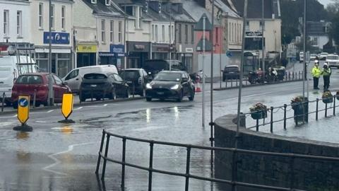 A rain-soaked town street with standing water flooding the roadside, cars driving through wet conditions, and traffic officers visible in the distance near a row of shops and houses under grey skies.
