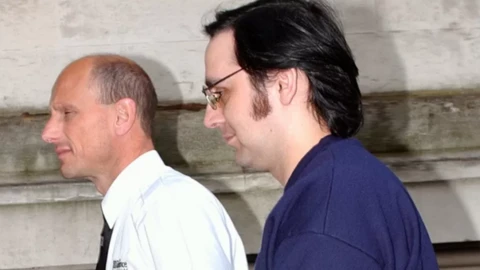 Jeffrey Gafoor being led into court by a prison guard. He has swept back dark hair, large sideburns and glasses and is wearing a dark blue jumper