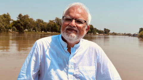 Shakil Salam sitting on a boat, on a river. He had short white hair, is wearing glasses, has a white beard and is wearing a white top.