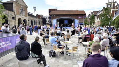A stage with a band performing on it. There are white blocks spelling "BBC" on the stage. There is a crowd of people watching, and most are sitting at tables and chairs.