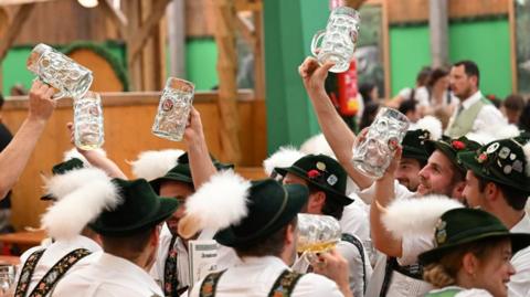 Multiple men and one woman dressed in traditional Oktoberfest outfits - Lederhosen: part pants and part shorts, held up by suspenders. They also have dark green hats on with a furry tail and badges on the top of the hat. Some of the men are holding up large empty beer glasses.