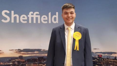 An image of a man with short mousey-dark hair. He is wearing a grey blazer with a white shirt and pale yellow tie, and has a bright yellow rosette on his left lapel. He is standing in front of a photograph image with the world Sheffield in white text in the left hand of the picture.