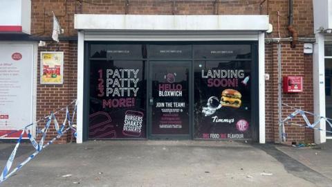 The front of the takeaway can be seen across the wide pavement outside. There are promotional posters stuck to the windows that tell people the burgers it will be producing are "landing soon". They also say "Hello Bloxwich". In the foreground police tape can be seen tied to a lamp-post.
