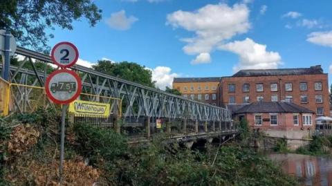 A steel footbridge over a river with some historic, brown-brick buildings in the background.