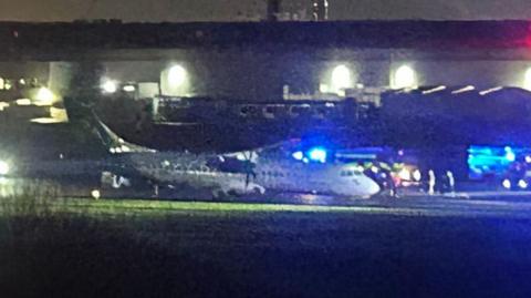 An Aer Lingus plane on a runway at nighttime surrounded by the blue lights of the emergency services. The plane's front wheel appears to have collapsed and its nose is resting on the airport tarmac