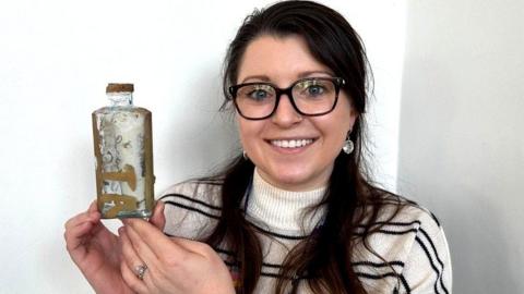 Abby Pendlebury has glasses and long brown hair. She is smiling and holding the bottle up to her face. She has ear piercings and a nose piercing.