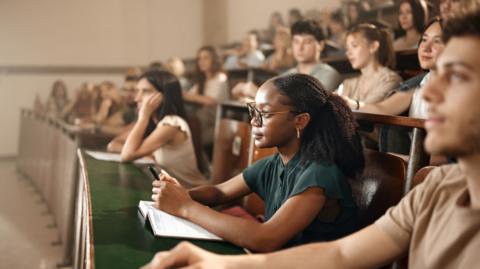 A young black woman wearing glasses is sitting at the front of a lecture hall, listening attentively - other students are visible around her