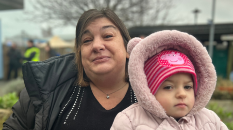 Heidi is wearing a black coat and shirt, and is holding her daughter who is in a pink coat and hat. 