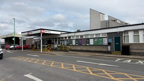 The exterior of Bletchley station which has a single-storey functional ticket office and waiting room, with a ramp and yellow handrail. There is a wooden portico with a station sign around the top. There are posters along the wall, and a single brown door to the right. There is doubled-door entrance under the portico. There is a strip of yellow "no waiting" paint on the roadway, which has white direction arrows on both sides.