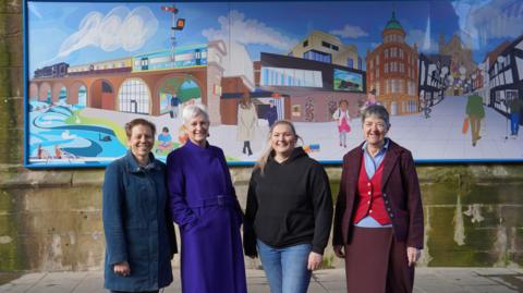 A group of women stand in front of a large rectangular mural. The mural features colourful depictions of local landmarks and buildings.