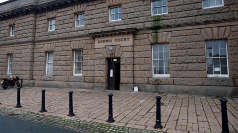 The exterior of Chester Crown Court, a large stone building with six black bollards outside, and a motorbike parked to the left of the image. Above a black door, is a light and a sign reading 'CROWN COURT'.