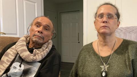 A man in a black t-shirt with cream and brown scarf and a woman wearing glasses in a green top with a star on it and a necklace