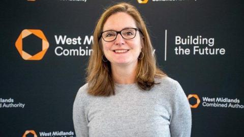 Laura Shoaf stood against a WMCA backdrop, wearing a grey top and black glasses. She is smiling. 
