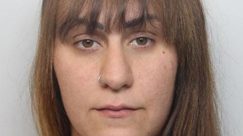 A mugshot photo of a woman with long brown hair stood against a grey background. She has a nose ring piercing.