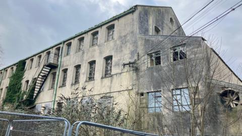 A run-down three-story building, with trees crawling up the wall and put out windows. The sky behind is grey and there is a metal fence around the perimeter