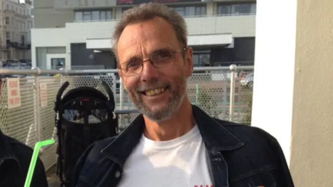 A man with brown hair, a beard and glasses wearing a white tshirt and black jacket standing on a balcony with a metal fence behind him