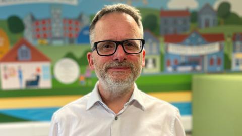 Phil Kloer looks at the camera. He has short grey hair and a short grey beard. He is wearing glasses and a white shirt and is standing in front of a mural with houses, the background is slightly out of focus. 