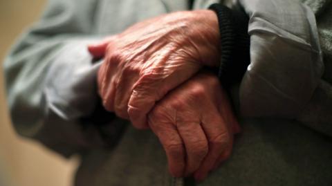 A stock image of the hands of an elderly person who is wearing a jacket and holding their hands one on top of the other across their body.