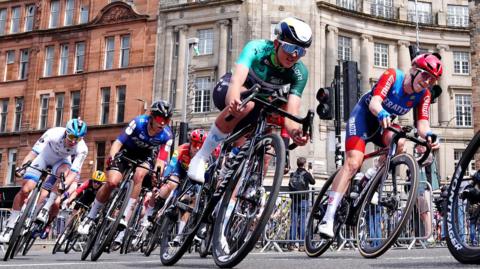 A number of cyclists wearing different coloured outfits, ride past during a race held on the streets of a city.