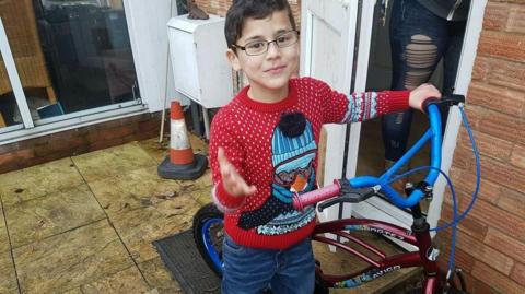 A boy stands on a garden patio with a bike. He has dark brown hair and glasses, and he is wearing a red Christmas jumper with a penguin on it. The bike is dark red with blue handlebars.
